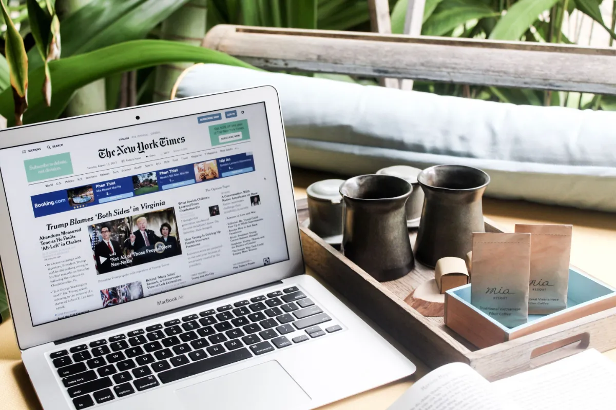 A laptop displaying a newspaper front page on a tray beside coffee cups and a plant