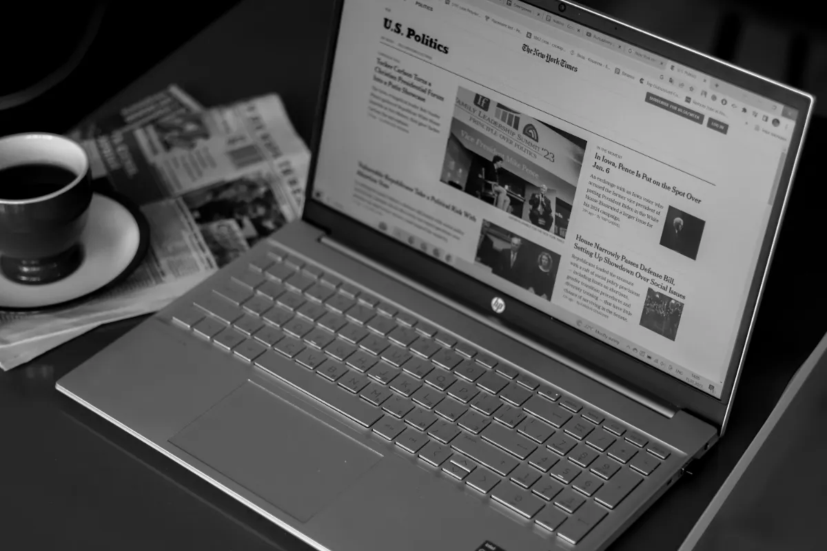 A black and white photograph of an open laptop showing a news site beside a coffee cup and folded newspapers